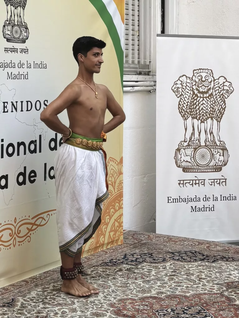 francisco valiente, bharatanatyam teacher and performer in madrid is dancing an item at the embassy of india in madrid. He is wearing colorful dress with the indian colors, orange white and green and posing with a royal pose in front of banners announcing The indian independence day.
