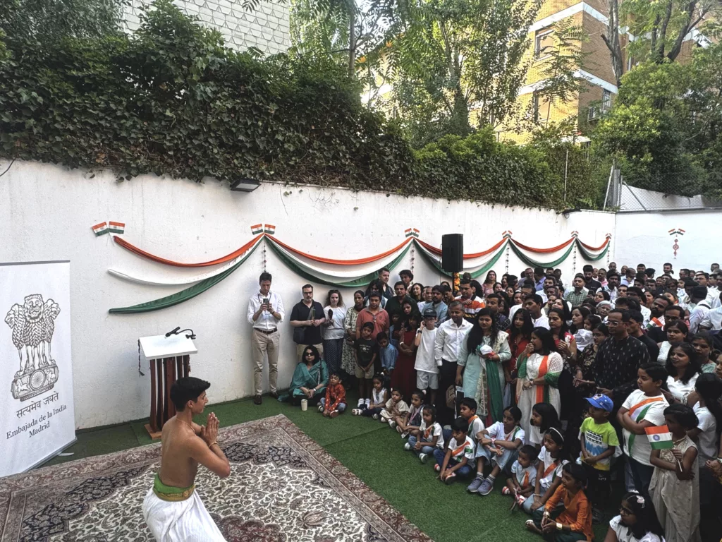 francisco valiente, bharatanatyam teacher and performer in madrid is dancing an item at the embassy of india in madrid. He is wearing colorful dress with the indian colors, orange white and green and posing with a royal pose in front of banners announcing The indian independence day. The audience is standing in front of francisco valiente and admiring his performance